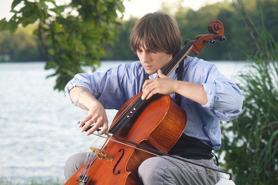 Elliott playing the cello outdoors by a lake, eyes on the strings, in a blue shirt and chef's apron