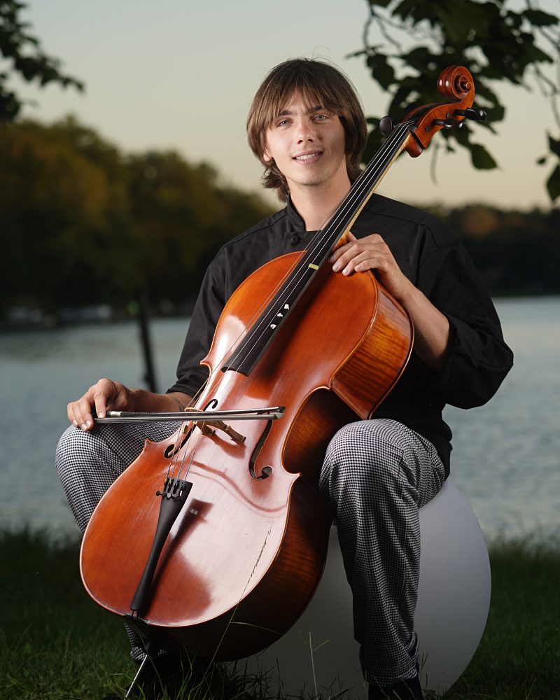 Chef Elliott in a black chef coat, smiling, holding his cello by a lake at dusk